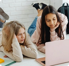 Két lány fekszik a padlón, és együtt használják a laptopot, ami az online tanulást, a csapatmunkát és a diákok közötti együttműködést szimbolizálja