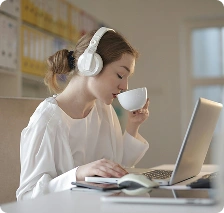Fejhallgatót viselő nő, aki laptopot használ és kávét iszik, szimbolizálva az online tanulást, a távmunkát és a koncentrált munkavégzést