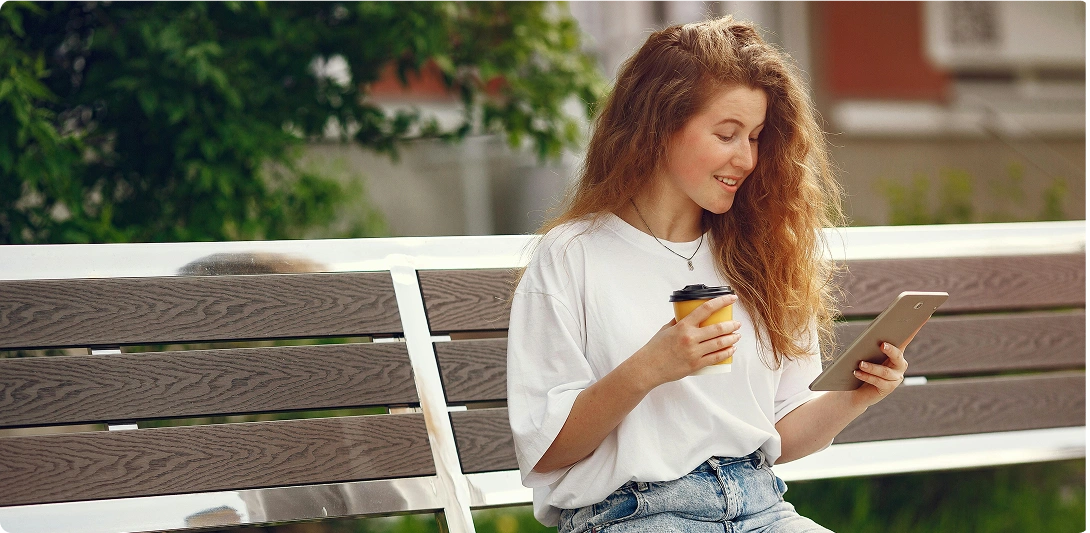 egy padon ülő fiatal nő, aki táblagépet használ és kávét tart a kezében; a kép a digitális életmódot, az internetes böngészést és a nyugodt mindennapokat ábrázolja