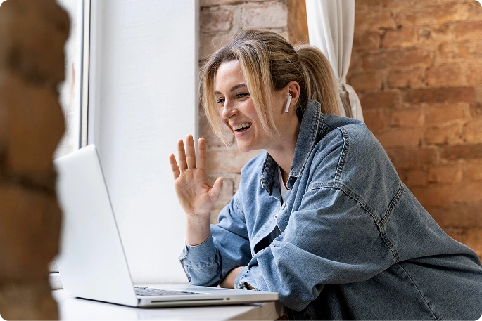 Egy nő mosolyogva integet egy laptopon zajló videohívás közben, szimbolizálva az online kommunikációt, a távmunkát és a virtuális találkozókat