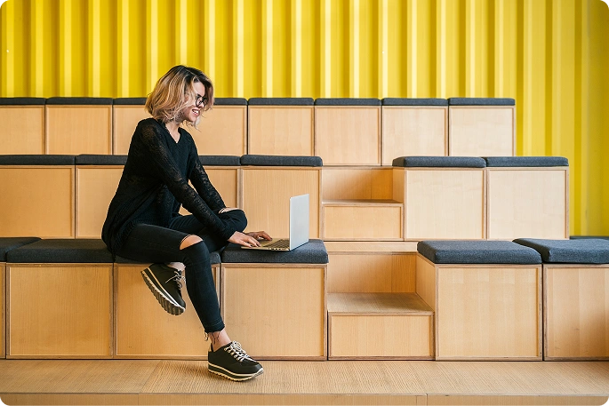 egy fiatal nő, aki laptopjával egy modern irodai környezetben ül, szimbolizálva a távmunkát, a kreativitást és a rugalmas tanulási környezetet