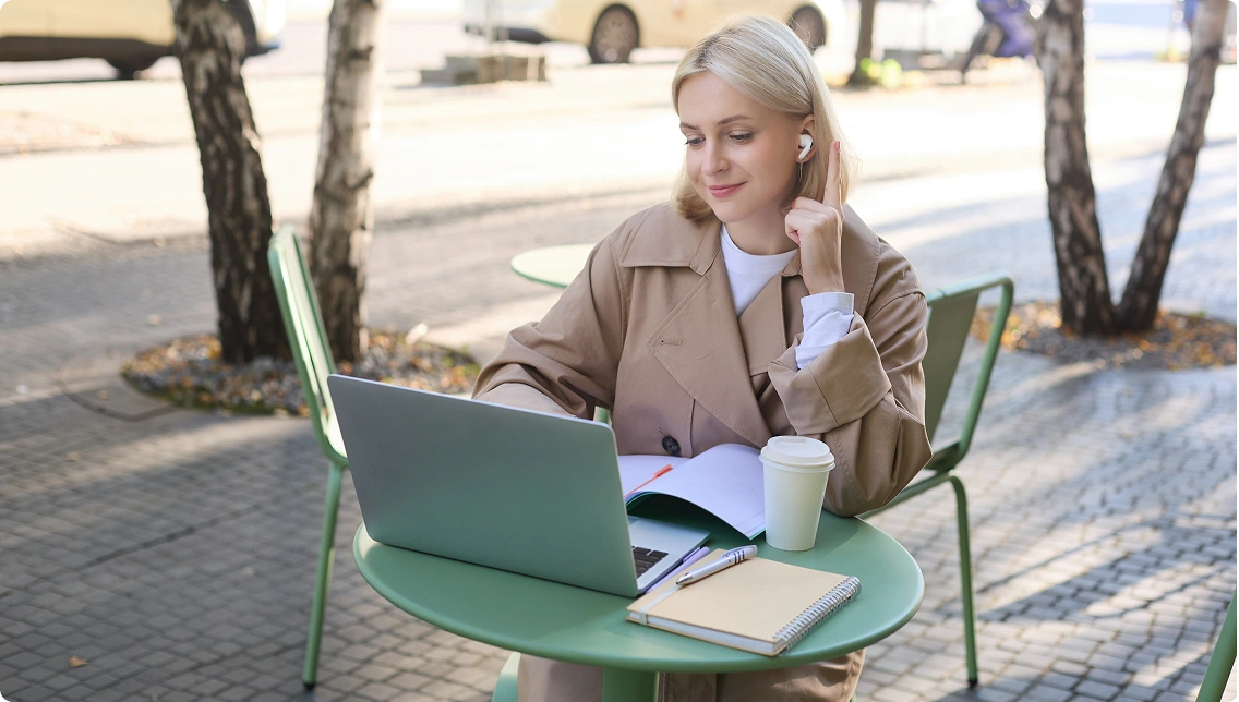 egy nő, aki egy kültéri asztalnál ülve laptopon dolgozik, kávéval és jegyzetfüzettel a kezében; a kép a távmunkát, az online tanulást és a rugalmas életmódot szimbolizálja