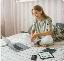 egy fiatal nő, aki az ágyon ülve laptopot használ és jegyzeteket ír, szimbolizálva az online tanulást, a távmunkát és a kényelmes otthoni tanulási környezetet